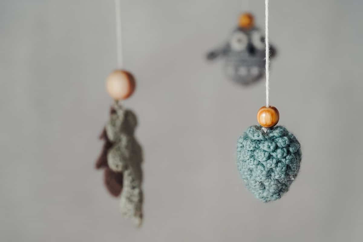 Small sage green crocheted ornaments with wooden beads hanging against a grey backdrop