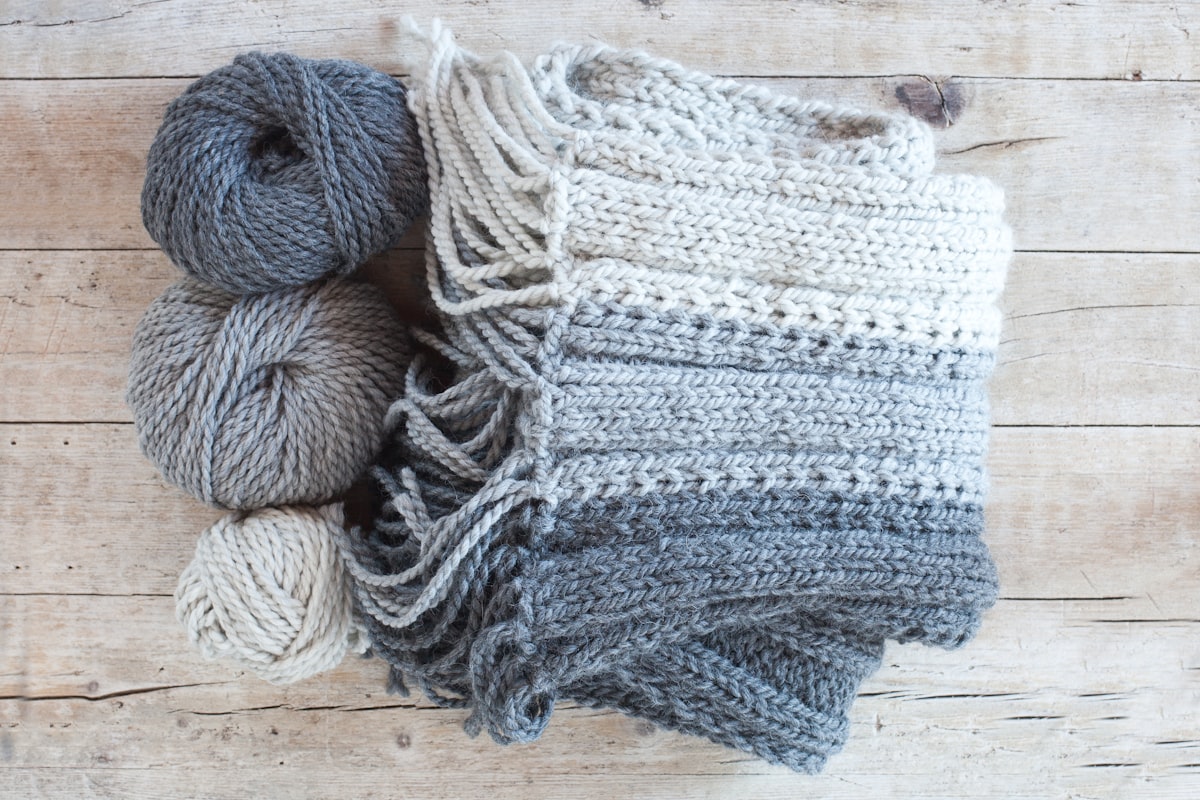 A grey and cream ombre crocheted scarf with matching yarn skeins on wood